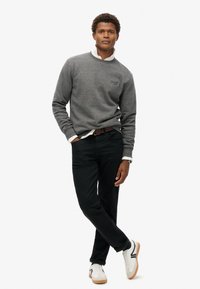 Suéter gris con cuello redondo, pantalones vaqueros negros y zapatillas blancas. Presenta puños acanalados y un logo sutil en el pecho. Diseño limpio.