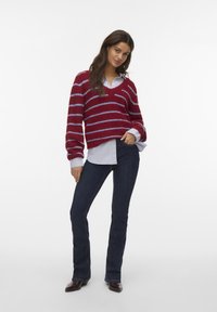 Frau, die einen rot-blauen gestreiften Pullover über einem hellblauen Hemd, dunkelblauen Jeans und braunen spitzen Schuhen trägt, selbstbewusst posiert.