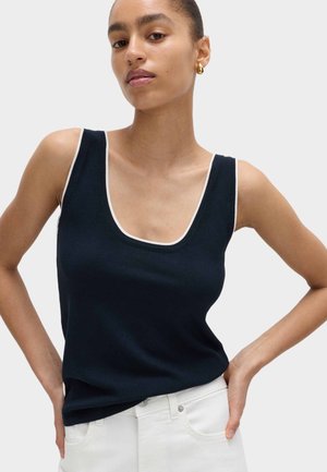 Kobieta ubrana w granatowy bezrękawnik z białą lamówką i białe spodnie, stojąca z rękami na biodrach na jednolitym tle.