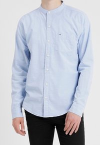 Light blue long-sleeve shirt with a mandarin collar, front-button closure, left chest pocket, and small logo detail. Smooth fabric texture.