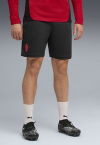 Svarte treningsshorts med en rød logo, kombinert med hvite sokker og svarte fotballsko. Stoffet er glatt og designet for komfort under sport.