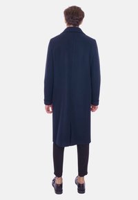 Cappotto lungo blu navy realizzato in lana, con taglio dritto, spacco posteriore e colletto a punta. Indossato sopra pantaloni slim neri e scarpe.