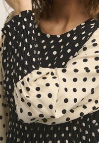 Långärmad klänning med svartvitt prickmönster, rynkade ärmslut och rund halsringning, bärs av en person med en arm höjd.