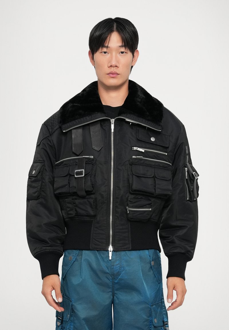 Chaqueta bomber negra con cuello de piel sintética, múltiples bolsillos frontales, detalles de cremallera y puños acanalados. Llevada sobre una camisa negra y pantalones azules.