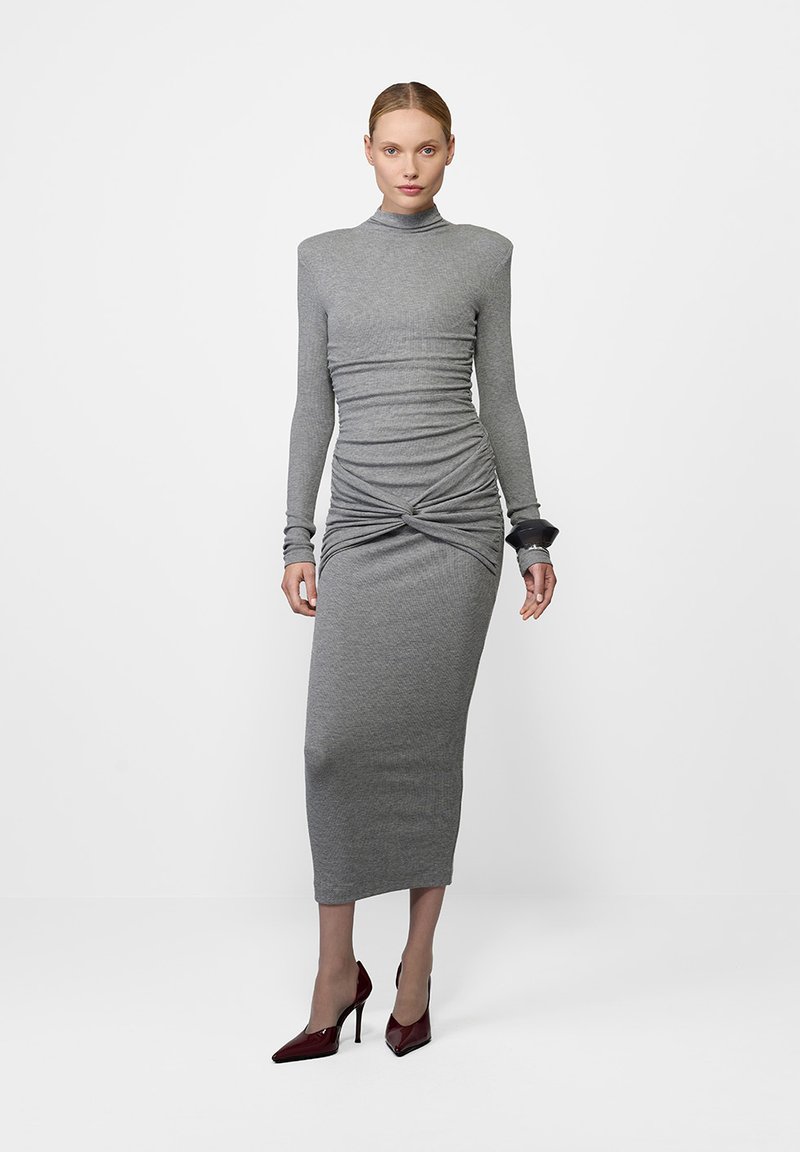 Halli soontega hall pikkade varrukate ja kogumisdetailidega vöökoht, kõrge kaelusega ning sääre keskpaigani ulatuva figuuri kehakujuga.