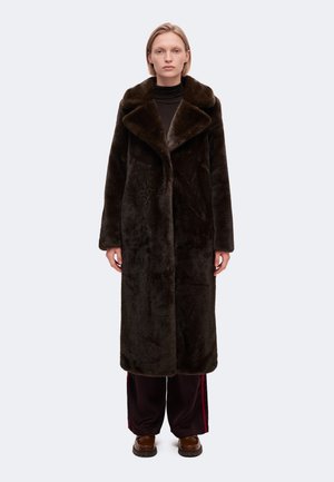 Un mannequin se tient devant un fond uni, vêtu d’un long manteau de fourrure brun foncé, d’un haut noir, d’un pantalon bordeaux et de chaussures marron.