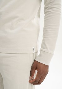 Helle beige Langarmbluse mit gerippten Bündchen und einem kleinen eingenähten Etikett mit der Aufschrift "MELA" am Saum, kombiniert mit passenden hellbeigen Hosen.