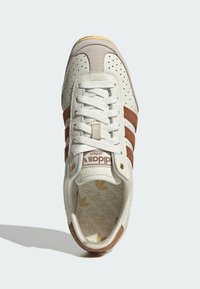 White leather sneaker with brown suede accents and perforated toe. Features white laces, gold hardware, and a patterned insole.