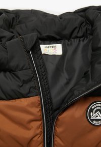 Czarna i brązowa izolowana kurtka z zapięciem na zamek, z okrągłą łatą z logo i miękką podszewką. Zaprojektowana dla dzieci z dopasowanym krojem.