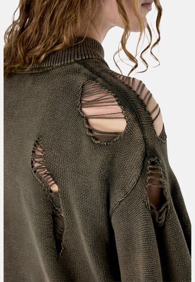 Kobieta o kręconych włosach, ubrana w ciemnozielony sweter z grubym splotem, z celowo dużymi dziurami i postrzępionymi brzegami na ramieniu i plecach.