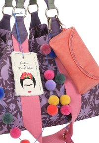 Paarse stoffen tas met bloemenpatroon, roze hengsels, kleurrijke pompons en een roze leren portemonnee. Inclusief een Frida Kahlo-label.