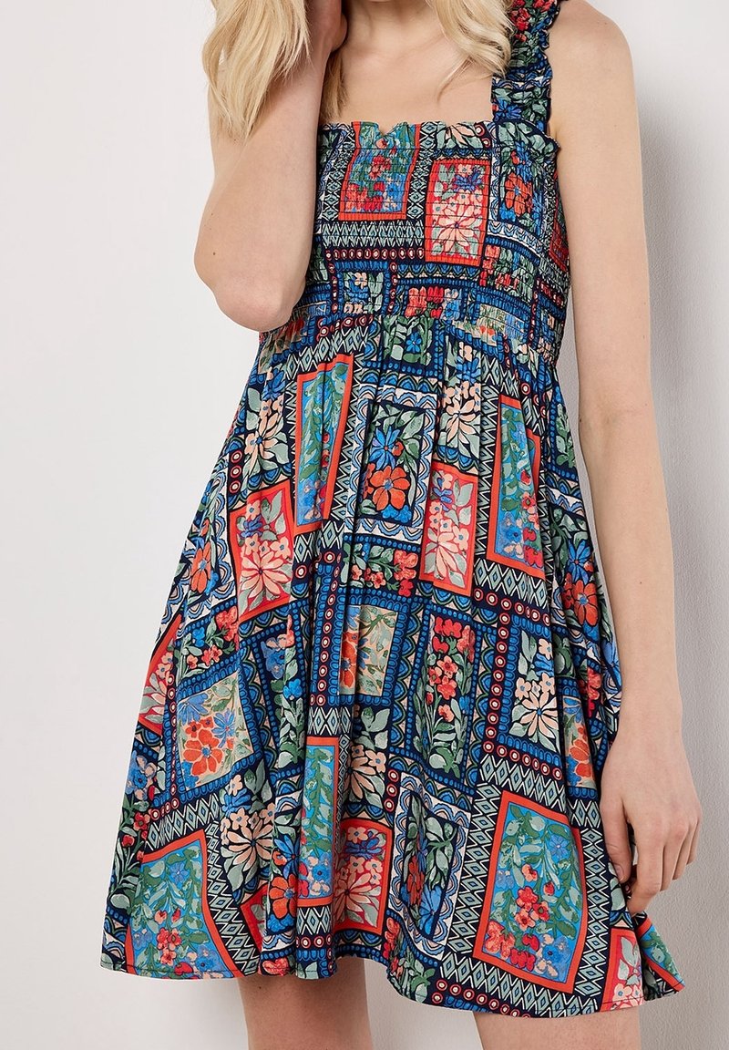 Femme portant une robe sans manches avec des motifs patchwork floraux et géométriques colorés dans des tons rouges, bleus, verts et beige.