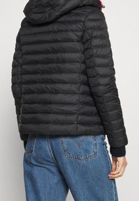 Giacca a vento nera con design a righe orizzontali e cappuccio. Il tessuto appare liscio e leggermente lucido. Abbinata a jeans di denim blu.