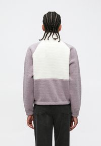 Veste polaire en lavande avec un panneau texturé blanc sur le haut du dos. Dotée d'un col montant et de poignets côtelés.