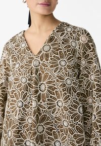 Top en tissu brun transparent avec un col en V, orné d'un motif de dentelle florale en crème. Accentué par des détails délicats et une coupe fluide.