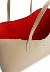 Beige tote bag met een gladde leren buitenkant, rode voering aan de binnenkant, twee bovenhandvatten en subtiele gouden branding aan de voorkant.