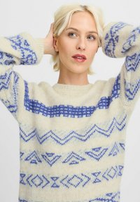Pulover din crem cu textură ușor evidentă, cu modele geometrice albastre și accente în zigzag, cu guler rotund și mâneci lungi, supradimensionate.