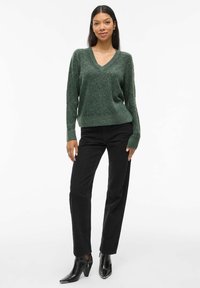 Pull vert en V en tissu texturé, associé à un jean noir taille haute et des bottes à talons brillantes noires. Le mannequin se tient avec assurance.