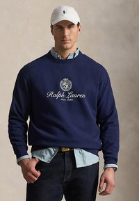 Man die een marineblauwe Ralph Lauren trui draagt over een lichtblauw overhemd met knopen, donkere spijkerbroek, witte pet en polshorloge, staand tegen een neutrale achtergrond.