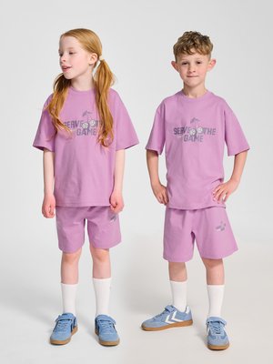 Deux enfants portant des t-shirts et shorts violets assortis "Serve the Game", avec des chaussettes blanches et des baskets bleues, debout sur un fond uni.