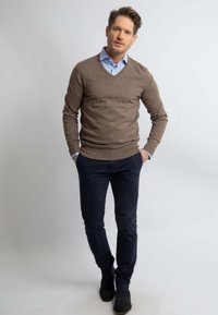 Bruine gebreide trui met een V-hals, gelaagd over een lichtblauwe overhemd met kraag. Donkerblauwe broek en zwarte schoenen maken de outfit af.