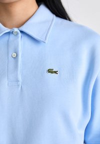 Šviesiai mėlyna polo marškinėliai su apykakle, turintys dvi mygtukus ir išsiuvinėtą žalią krokodilo logotipą kairėje krūtinės pusėje. Minkšta, tekstūruota medžiaga.