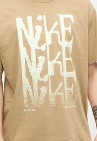 Μπεζ μπλούζα με μεγάλο, λευκό, ψηφιδωτό κείμενο "NIKE", μικρότερο κείμενο "NIKE INC." και "OREGON, USA" στο κάτω μέρος.