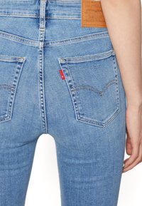 Jean en denim bleu clair à taille haute, avec des poches arrière ornées de surpiqûres arquées et un patch de marque en cuir marron à l'arrière.