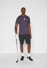 Lila pamut póló hímzett Nike logóval, fekete rövidnadrággal párosítva. Egy golfozó golfütőt tart, fekete tornacipőt és fehér zoknit viselve.