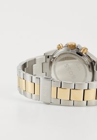 Orologio in acciaio inox con bracciale in tonalità argento e oro. Presenta tre pulsanti in oro e una cassa rotonda con fondo in vetro minerale.