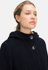 Marineblauwe wollen jas met een capuchon, voorzien van twee grote zwarte knopen en een zachte textuur, ontworpen met een strakke, minimalistische esthetiek.