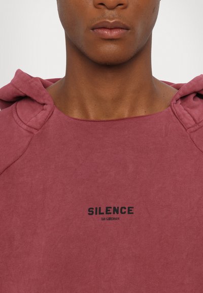 Sweatshirt com capuz na cor rosa suave, feita de algodão macio. Apresenta um decote largo, um corte oversized e a palavra "SILÊNCIO" impressa em preto no peito.