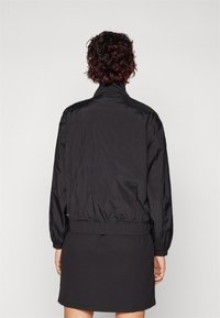 Veste bomber noire, légère, avec col montant, poignets et taille élastiques, présentant une texture lisse et un minimalisme dans les détails.