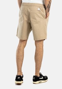 Pantalones cortos color caqui con cintura elástica, dos bolsillos traseros y dobladillo plano. Se llevan con zapatillas negras que tienen un logo blanco.