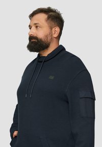 Sweat à capuche bleu foncé fabriqué en tissu texturé, avec cordons de serrage, poches latérales et une patch sur la poitrine gauche et la manche.