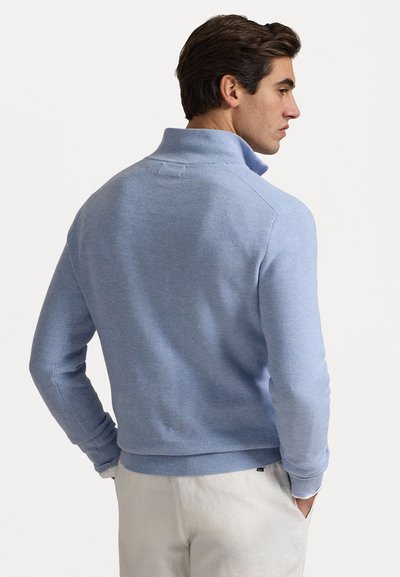 Pull en tricot bleu clair avec col montant, présentant un motif texturé et des manches côtelées, associé à un pantalon de couleur claire.