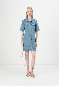 Vestido de camisa em denim, com mangas curtas, cor azul clara, fecho frontal com botões, dois bolsos no peito e um corte descontraído. Acentuado com detalhes dourados.