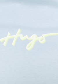 Světle modré tričko s hladkým povrchem, které se pyšní velkým bílým kurzivním logem "Hugo" s žlutým okrajem.