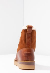 Botín de ante marrón hasta el tobillo con un tacón de cuero marrón claro, borde de borreguillo en la parte superior y suela de goma. Incluye una lengüeta para facilitar su uso.