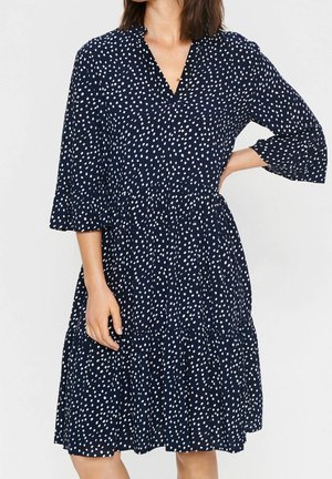 Femme portant une robe bleu marine longueur genoux à pois blancs et manches trois-quarts, debout avec une main sur la hanche.
