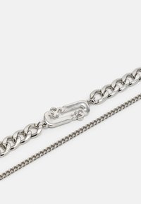 Colar de corrente prateada com um elos maior entrelaçado, com detalhes gravados, textura suave e uma corrente mais fina em contraste.