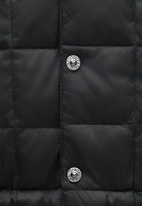 Sort puffervest med quiltet design, der har en shinny, glat tekstur og to metalsnapknapper med branddetaljer.