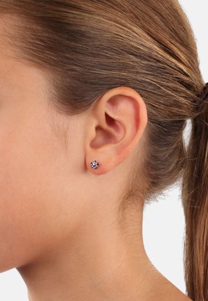 Pendientes de botón con un diseño floral y piedras preciosas moradas engastadas en metal, llevados en la oreja de una persona. El cabello está recogido. La piel parece suave.