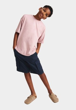 Junge trägt ein hellrosa übergroßes T-Shirt, marineblaue Cargo-Shorts und beige Slip-On-Schuhe, steht mit den Händen in den Taschen und geneigtem Kopf.