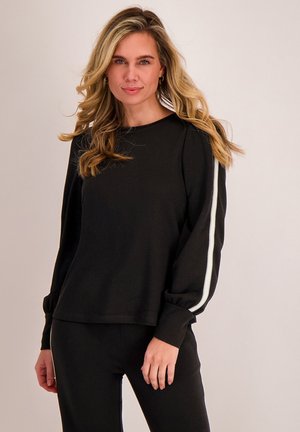 Vrouw met lang blond haar, gekleed in een zwart langarmig shirt en een broek met witte strepen langs de mouwen en benen, staand tegen een effen achtergrond.
