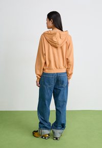 Avkortet, brent oransje hettegenser med snorhette, kombinert med løse blå jeans og svarte og gule sneakers. Grønn gulvbakgrunn.
