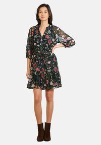 Kobieta ubrana w ciemną sukienkę w kwiaty do kolan z bufkami, z paskiem w talii i brązowymi botkami sięgającymi kostek, stojąca z jedną ręką na biodrze.