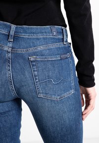 Záběr zblízka na osobu v modrých džínech a černém topu s dlouhým rukávem, zaměřující se na detail zadní kapsy a ruku, která odpočívá po boku.