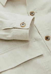 Beige katoenen shirt met een opgevouwen manchet met twee ronde, bruine knopen en een borstzak, met een gladde textuur en subtiele stiksels.