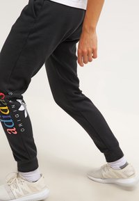 Osoba v černých joggerách Adidas Originals s barevným logem a bílými teniskami, stojící s jednou nohou vpřed na světlém pozadí.